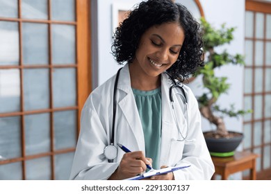 Happy biracial female doctor holding clipboard and taking notes at doctor's office. Hospital, medicine and healthcare. - Powered by Shutterstock