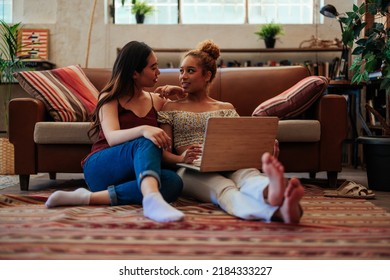 Happy Biracial Gay Women Using Laptop While Sitting On The Floor At Home, Leaning On Sofa.