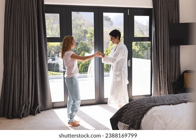 Happy biracial lesbian couple holding hands dancing in sunny bedroom at home, copy space. Summer, gay, fun, relationship, togetherness, domestic life and lifestyle, unaltered. - Powered by Shutterstock