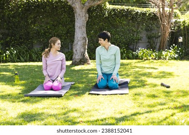 Happy biracial lesbian couple practicing yoga kneeling on mats in sunny garden, copy space. Summer, gay, relationship, togetherness, yoga, fitness and healthy lifestyle, unaltered. - Powered by Shutterstock