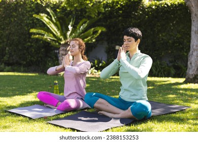 Happy biracial lesbian couple practicing yoga meditation sitting on mats in sunny garden. Summer, gay, relationship, togetherness, yoga, fitness and healthy lifestyle, unaltered. - Powered by Shutterstock