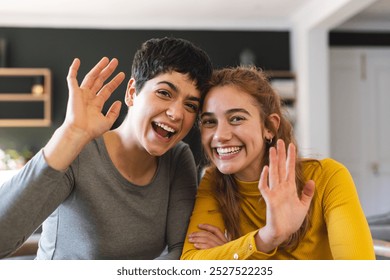 Happy biracial lesbian couple sitting at home having video call, waving. Communication, technology, gay, relationship, togetherness, domestic life and lifestyle, unaltered. - Powered by Shutterstock