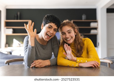 Happy biracial lesbian couple sitting at table having video call, waving. Communication, technology, gay, relationship, togetherness, domestic life and lifestyle, unaltered. - Powered by Shutterstock