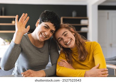 Happy biracial lesbian couple sitting at home having video call, waving. Communication, technology, gay, relationship, togetherness, domestic life and lifestyle, unaltered. - Powered by Shutterstock