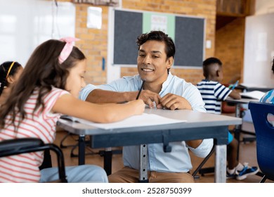 Happy biracial male teacher with girl in wheelchair in class at elementary school. School, learning, childhood, disability, inclusivity, teaching and education, unaltered. - Powered by Shutterstock