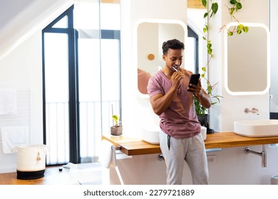 Happy biracial man brushing teeth and using smartphone in bathroom. Lifestyle, self care, communication and domestic life, unaltered.