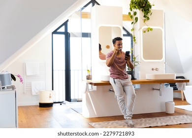 Happy biracial man brushing teeth and using smartphone in bathroom. Lifestyle, self care, communication and domestic life, unaltered. - Powered by Shutterstock