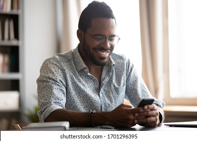 Happy biracial man in glasses sit at desk have fun texting messaging using wireless internet on smartphone gadget, smiling African American male in spectacles laugh watch funny video on cellphone - Powered by Shutterstock