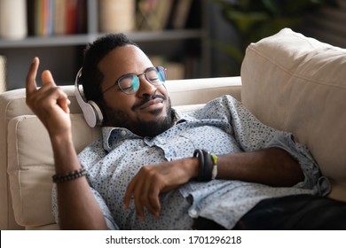 Happy Biracial Millennial Man In Wireless Headphones Relax On Couch At Home Listen To Music, Overjoyed African American Male Rest On Sofa In Living Room Enjoy Good Quality Sound In Modern Earphones