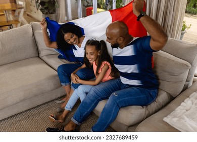 Happy biracial parents and daughter sitting on couch watching tv holding french flag and smiling. Family, sport, patriotism, celebration, togetherness, lifestyle and domestic life, unaltered. - Powered by Shutterstock