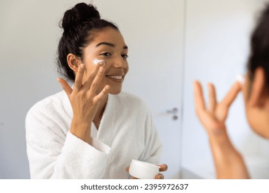 Happy biracial woman in bathrobe applying cream on her face in sunny bathroom. Lifestyle, self care, beauty, skin and domestic life, unaltered. - Powered by Shutterstock