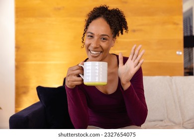 Happy biracial woman with cup of coffee making video call and waving in living room. Lifestyle, communication and domestic life, unaltered. - Powered by Shutterstock