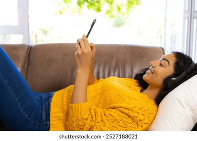 Happy biracial woman with headphones using smartphone lying on sofa at home. Lifestyle, free time, communication and domestic life, unaltered. - Powered by Shutterstock