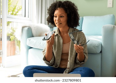 Happy biracial woman using laptop and talking on smartphone sitting on floor in bedroom. Lifestyle, communication and domestic life, unaltered. - Powered by Shutterstock