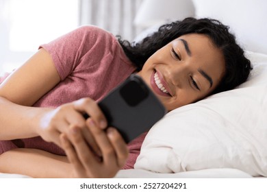 Happy biracial woman using smartphone lying on bed at sunny home. Lifestyle, free time, communication and domestic life, unaltered. - Powered by Shutterstock