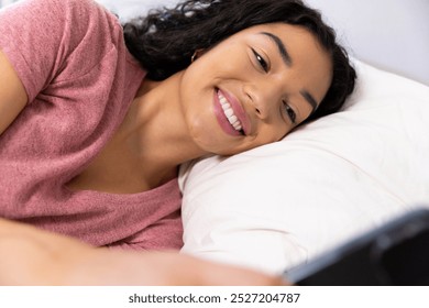 Happy biracial woman using smartphone lying on bed at sunny home. Lifestyle, free time, communication and domestic life, unaltered. - Powered by Shutterstock