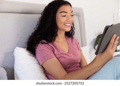 Happy biracial woman using tablet sitting on bed at sunny home. Lifestyle, free time, communication and domestic life, unaltered. - Powered by Shutterstock