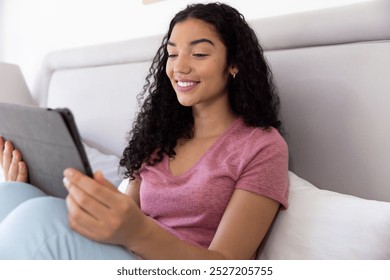 Happy biracial woman using tablet sitting on bed at sunny home. Lifestyle, free time, communication and domestic life, unaltered. - Powered by Shutterstock