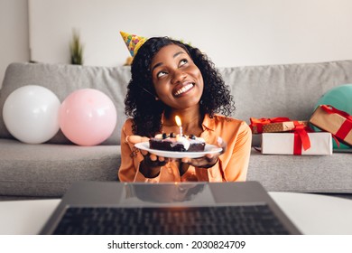 Happy Birthday. African American Female Making Wish Holding B-Day Cake In Front Of Laptop Computer Having Online Party Via Video Call At Home. Remote Holiday Celebration