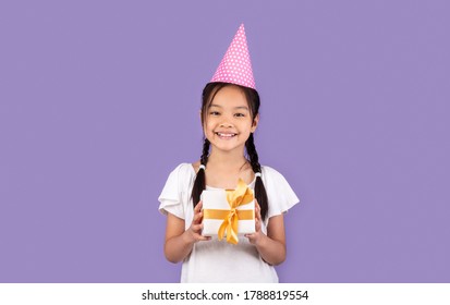 Happy Birthday. Asian Girl Holding B-Day Gift Box Wearing Pink Party Hat Posing Over Purple Background In Studio. Free Space