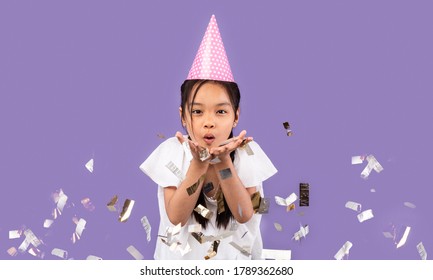 Happy Birthday. Chinese Kid Girl Blowing Silver Confetti At Camera Celebrating B-Day Wearing Pink Party Hat Over Purple Studio Background.