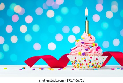 Happy Birthday Cupcake. Tasty Cupcakes With Pink Cream Icing And Colored Sprinkles. Burning Candle In A Cake. Sweet Delicious Dessert On White Wooden Table And Blue Background With Bokeh. Copy Space.