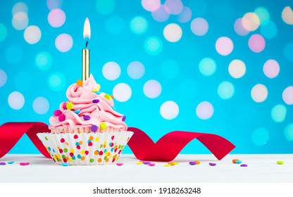 Happy Birthday Cupcake. Tasty Cupcakes With Pink Cream Icing And Colored Sprinkles. Burning Candle In A Cake. Sweet Delicious Dessert On White Wooden Table And Blue Background With Bokeh. Copy Space.