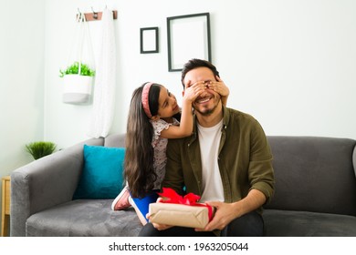 Happy Birthday, Dad! Hispanic Little Daughter Covering Her Father's Eyes To Give Him A Surprise Gift For His Birthday In The Living Room