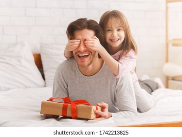 Happy birthday daddy. Preschool happy girl greeting her beloved young dad with birthday, covering his eyes, giving gift box, bedroom interior, free space - Powered by Shutterstock