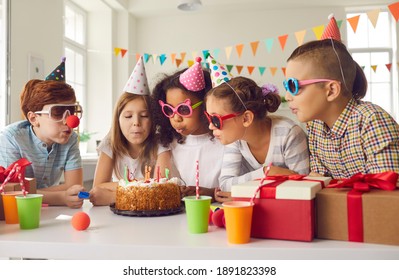 Happy Birthday. Group of diverse little children in paper hats and party sunglasses all together blowing candles on cake standing at festive table during fun celebration at home