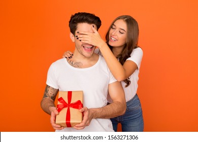 Happy Birthday, Honey! Woman Closing Husband's Eyes, Holding Gift For Him Over Orange Background