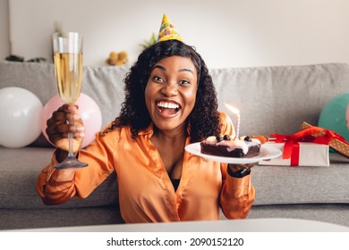 Happy Birthday. Joyful Black Lady Holding B-Day Cake With Candle And Glass Of Wine Smiling To Camera Posing Wearing Festive Hat, Celebrating Holiday Online Via Video Call At Home