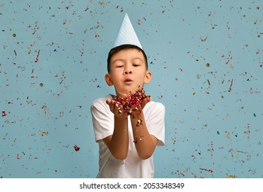 Happy Birthday. Little Asian Boy Wearing Party Hat Blowing Colorful Confetti At Camera, Cheerful Korean Male Child Enjoying Celebrating B-Day, Posing Over Blue Studio Background, Copy Space