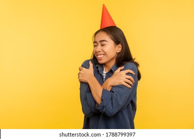 Happy Birthday To Me! Portrait Of Girl In Denim Shirt And With Funny Party Cone On Head Embracing Herself, Celebrating Successful Event Alone But Happy. Studio Shot Isolated On Yellow Background