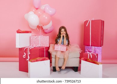 Happy birthday mood of cute little girl in tulle skirt smiling to camera with present on pink background. Celebrating surround big colorful giftboxes, balloons of fashionable young princess