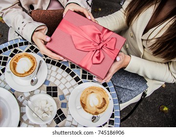 Happy Birthday! Top View Of Beautiful Woman Giving Present To Best Friend While Sitting In French Vintage Cafe. Receiving Gift From Friend. Birthday Celebration In Restaurant. Women Holding Gift Box