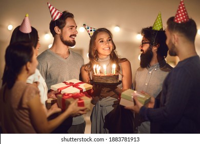 Happy Birthday To You. Smiling Young Woman In Her Twenties Holding Her Birthday Cake With Lit Candles And Thanking Her Friends For Surprise And Presents At Evening Party At Home