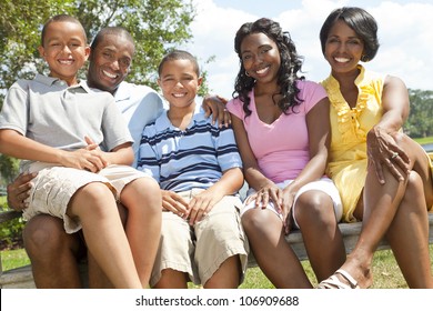 A Happy Black African American Family Of Two Parents And Three Children, Two Boys One Girl, Sitting Together Outside.