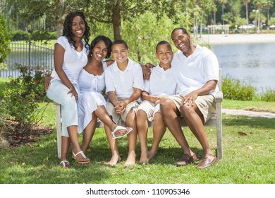 A Happy Black African American Family Of Two Parents And Three Children, Two Boys One Girl, Sitting Together Outside In White Clothes