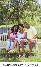 A Happy Black African American Family Of Two Parents And One Female Girl Child Sitting Together Outside On A Park Bench Laughing.