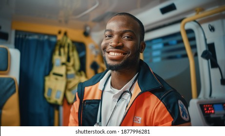 Happy Black African American Paramedic Smiles and Poses for Camera in an Ambulance Vehicle with an Injured Patient. Emergency Medical Technician is Cheerful at Work.