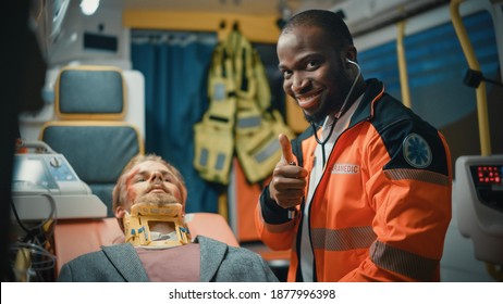 Happy Black African American Paramedic Poses For Camera And Gives A Thumbs Up In An Ambulance Vehicle With An Injured Male Patient. Emergency Medical Technician Is Using A Stethoscope.