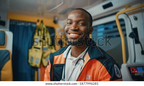 Happy Black African\
American Paramedic Smiles and Poses for Camera in an Ambulance\
Vehicle with an Injured Patient. Emergency Medical Technician is\
Cheerful at Work.