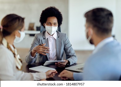 Happy Black Bank Manager With Face Mask Using Touchpad While Talking To Her Clients On A Meeting In The Office. 