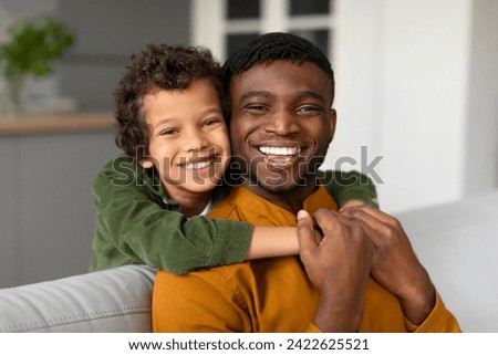 Happy black boy with curly hair joyfully hugs his father from behind, both sharing radiant smiles in comfortable home setting, man sitting on couch