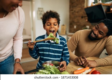 Happy black boy tasting salad while preparing healthy meal with parents in the kitchen. 