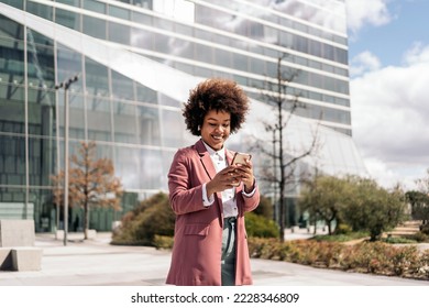 Happy black business woman with afro hair smiling and using her mobile phone outdoors in a business area. - Shutterstock ID 2228346809