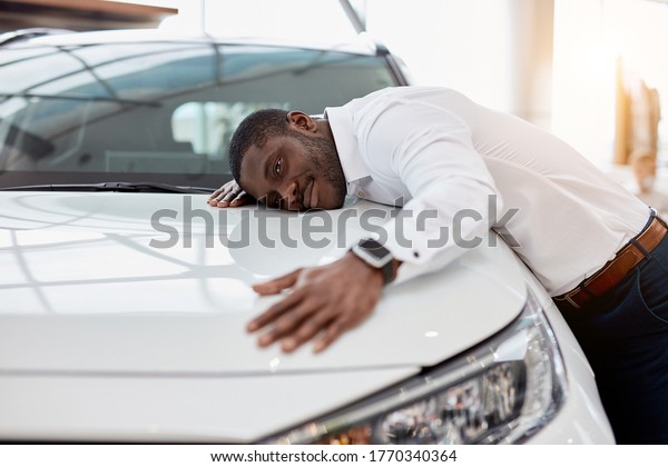 happy black businessman hug his\
new white luxurious car in dealership, he made successful\
purchase.