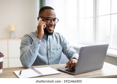 Happy Black Businessman Talking On Cellphone Having Phone Conversation Sitting At Workplace Working On Laptop Computer In Office. Man Chatting On Smartphone Indoor. Mobile Communication Concept