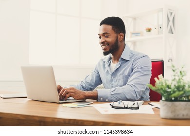 Happy Black Businessman Working On Laptop In Modern White Office Interior. Smiling Employee At Work With Computer, Copy Space